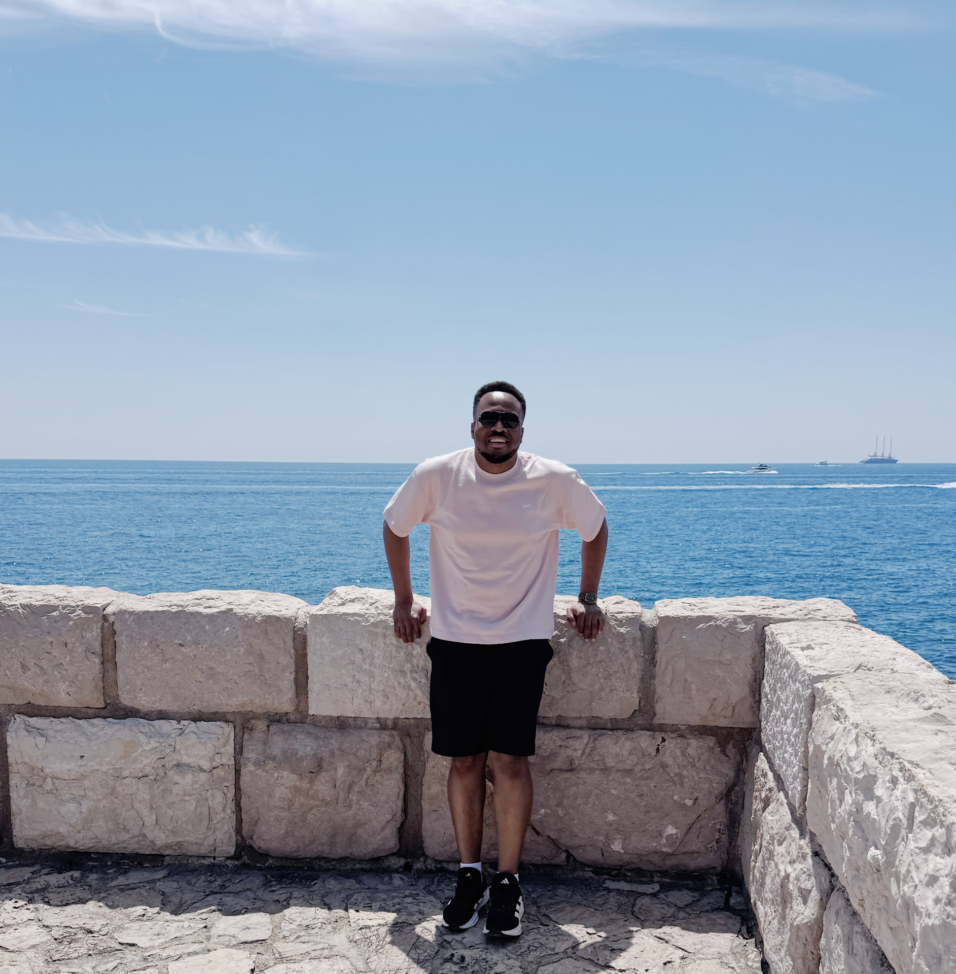 Uche Ojukwu resting against a stone sea wall on a bright day, open water behind him.