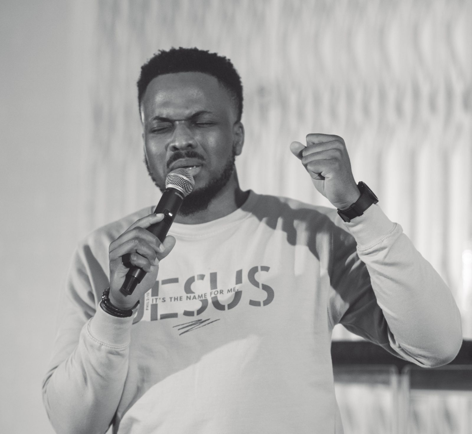 Uche Ojukwu preaching with a microphone, eyes closed mid-sentence, in black and white.