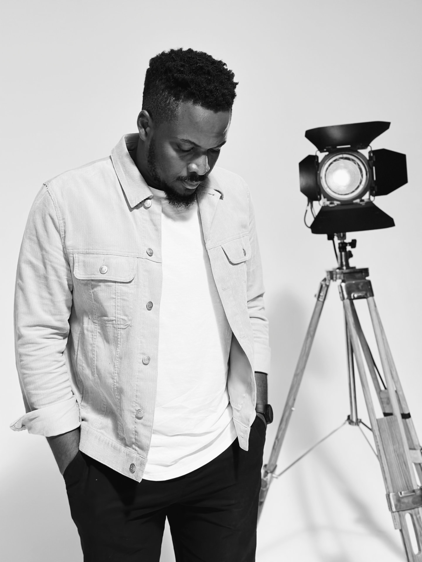 Uche Ojukwu looking down in thought beside a studio light, in black and white.